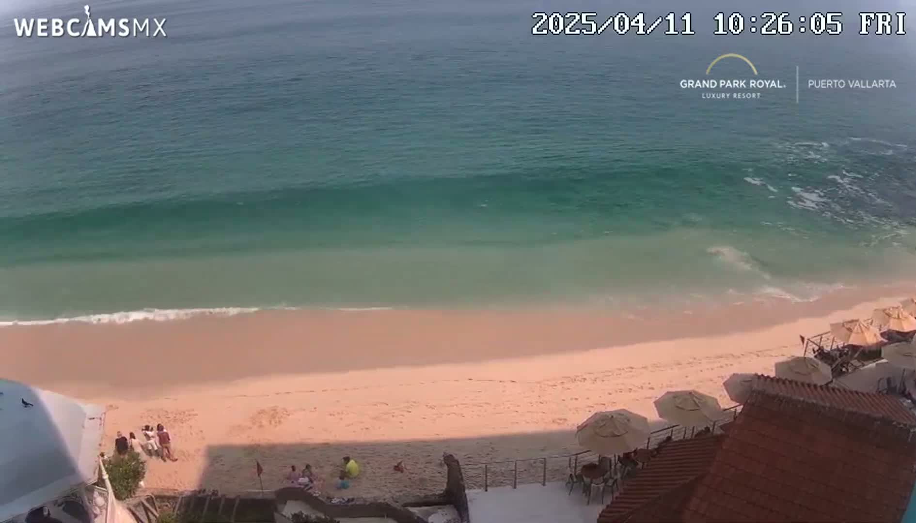 A sandy beach with a calm ocean and several people are visible near some beach umbrellas and buildings.