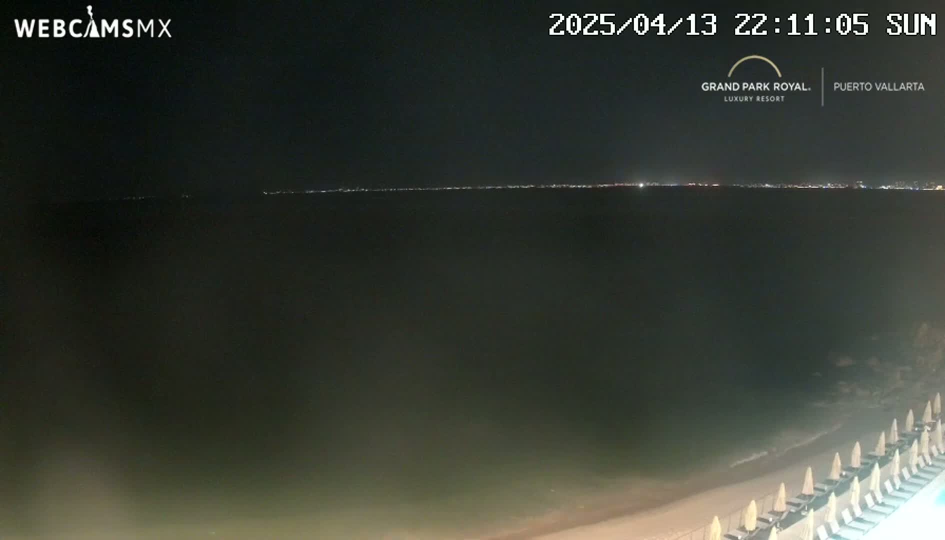 A nighttime beach scene shows a dark ocean with a distant city skyline illuminated, and beach chairs lined up along the sand.