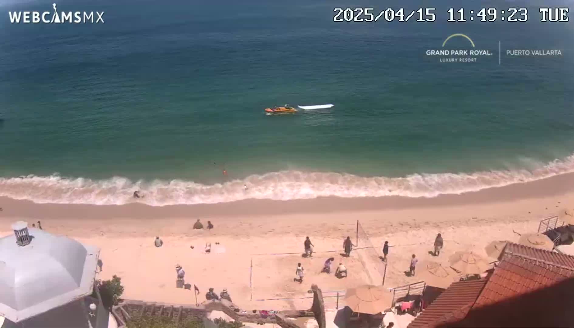 A sandy beach with several people on it, gentle waves lapping the shore, and a small boat on calm, clear ocean water.
