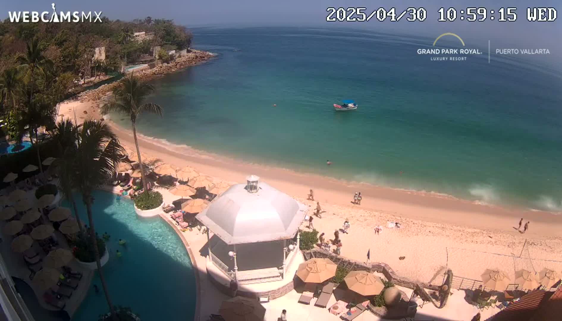An aerial view shows a beach with turquoise water, a resort pool, and a gazebo under a sunny sky.