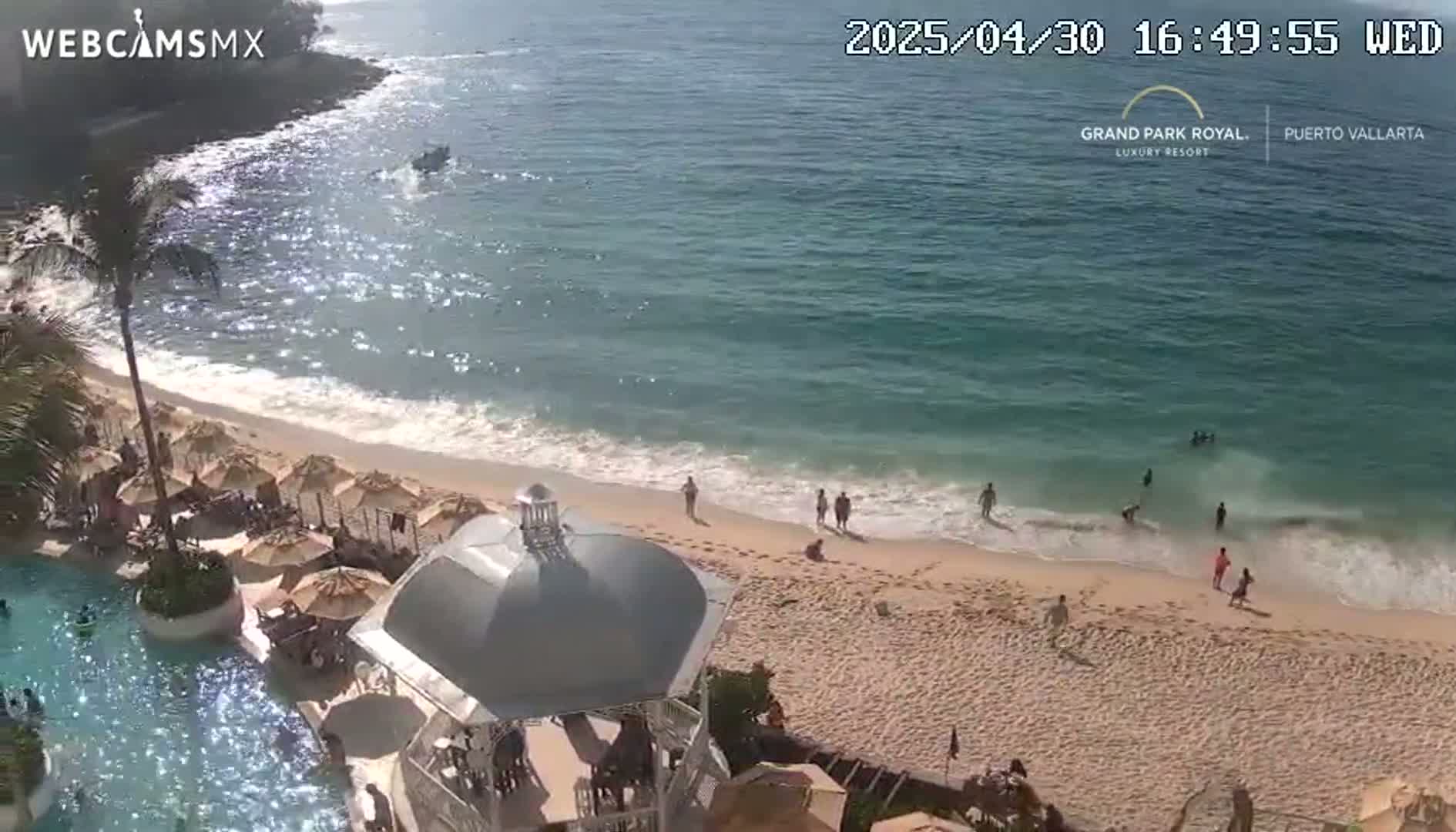 An aerial view shows a sandy beach with people swimming and relaxing near a resort pool and gazebo under sunny conditions.