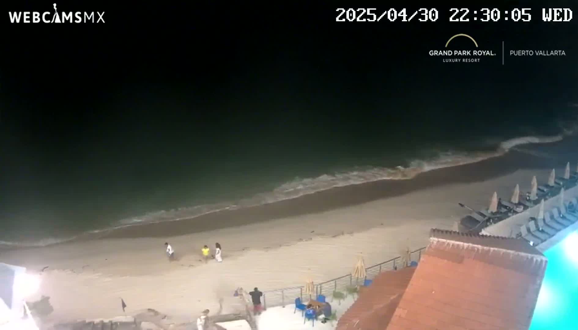 A dark, nighttime beach scene shows gentle waves lapping a sandy shore, with a few people on the beach near a resort.