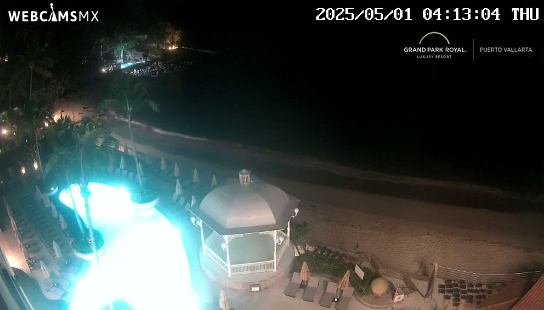 A nighttime view of a resort pool illuminated in teal, adjacent to a sandy beach and ocean with gentle waves under a dark sky.