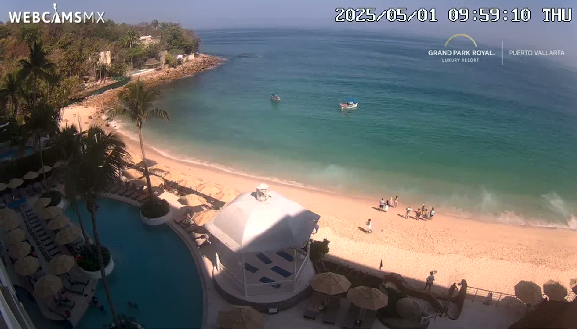 An aerial view shows a resort pool and beach on a sunny day, with calm turquoise water and a few people on the sandy shore.