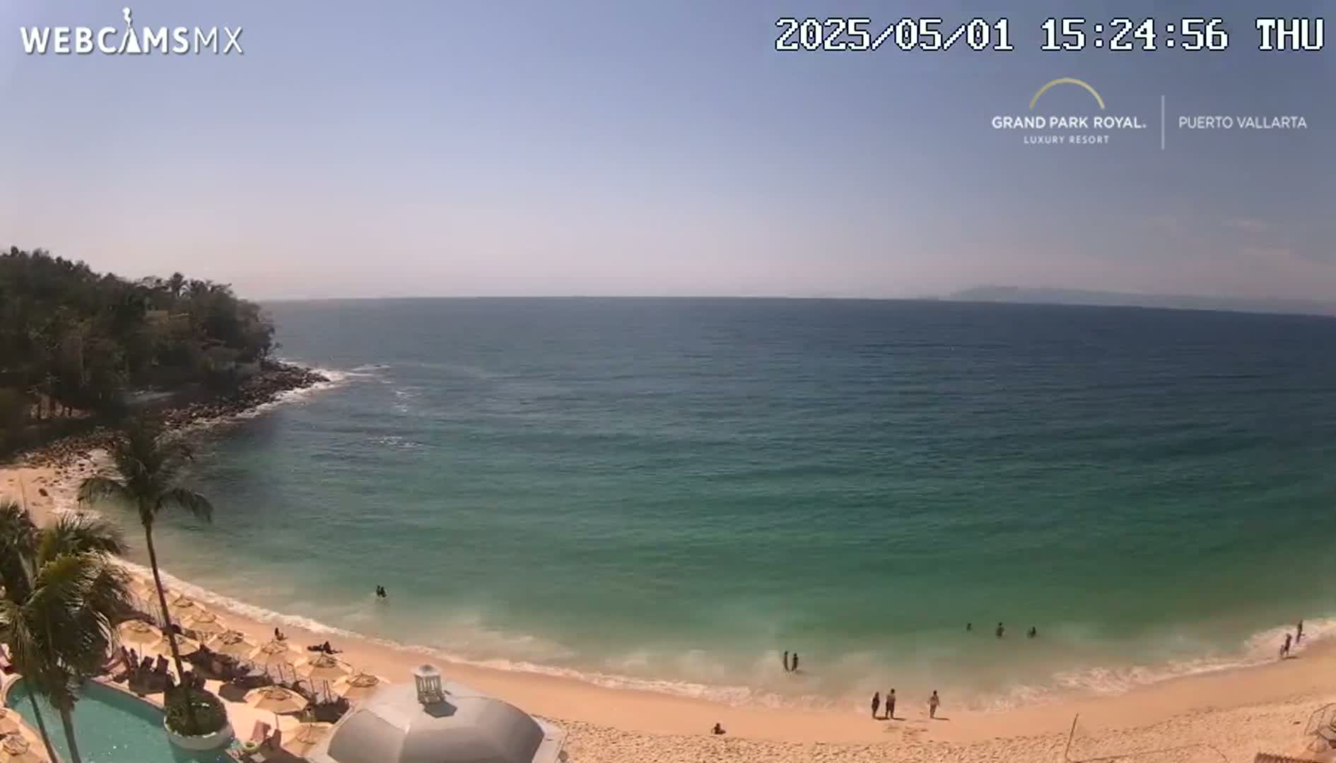 A sandy beach with calm, clear turquoise water and a few people swimming and sunbathing, under a clear blue sky.
