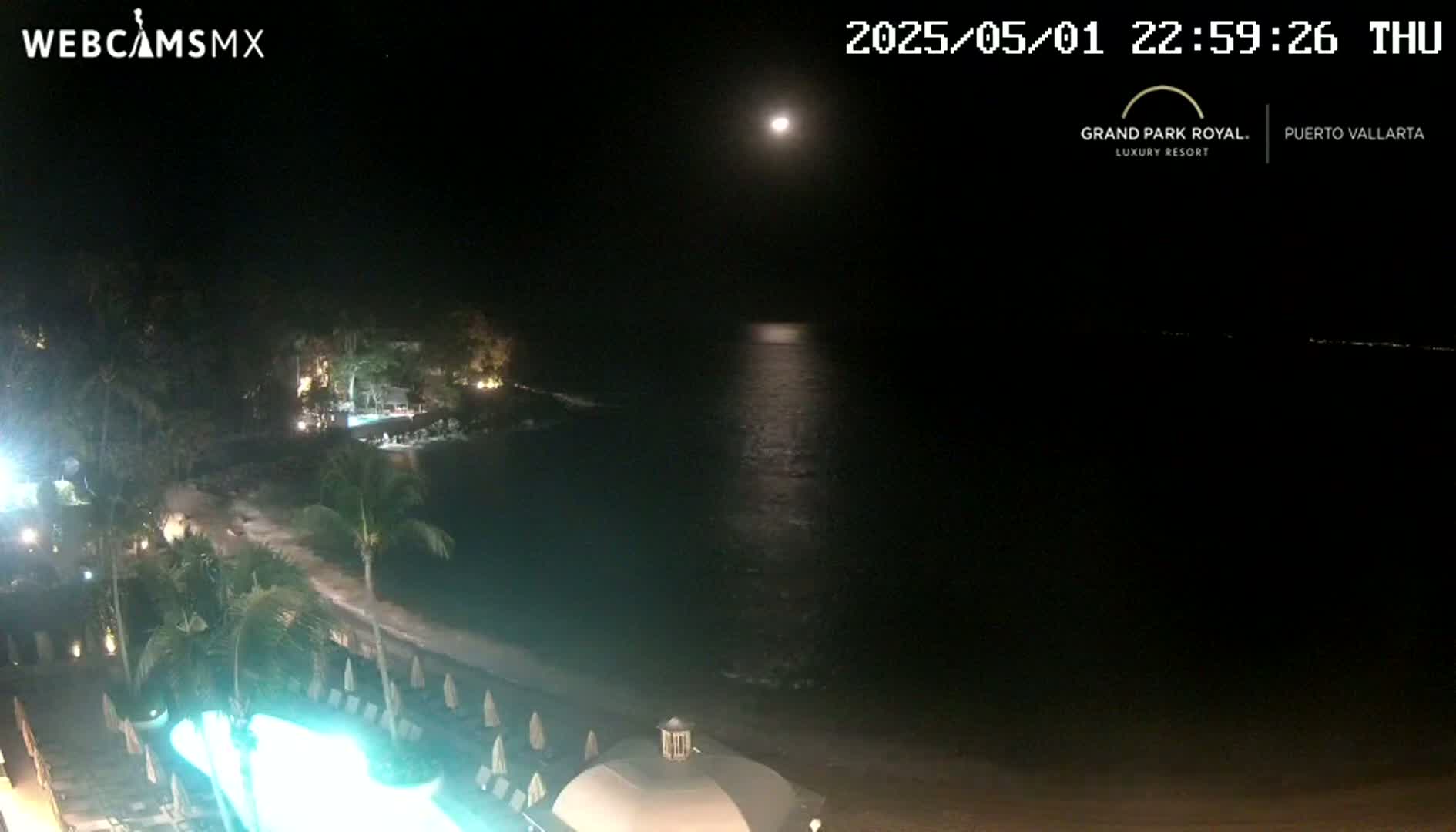 A nighttime view of a resort beach with a calm ocean under a bright moon.