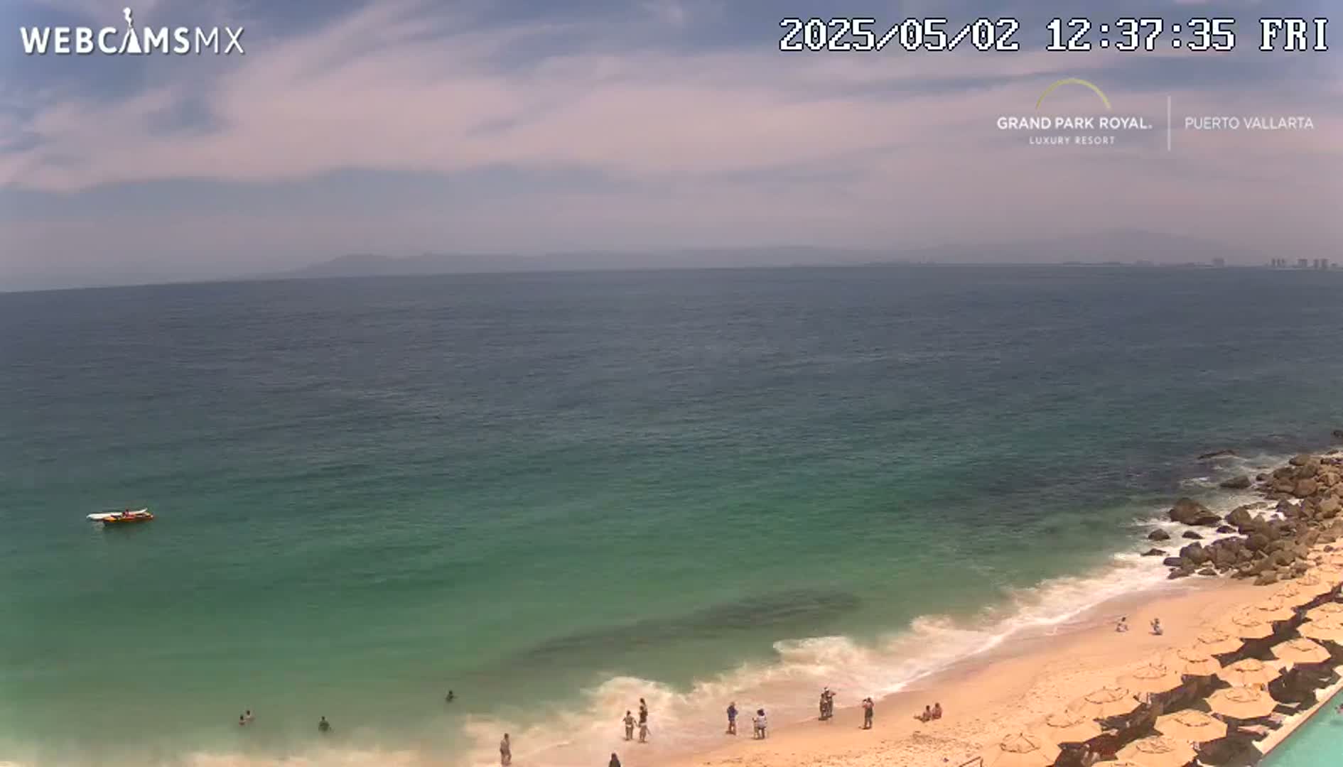 A partly cloudy day shows a calm ocean with a sandy beach and several people swimming and relaxing under beach umbrellas.