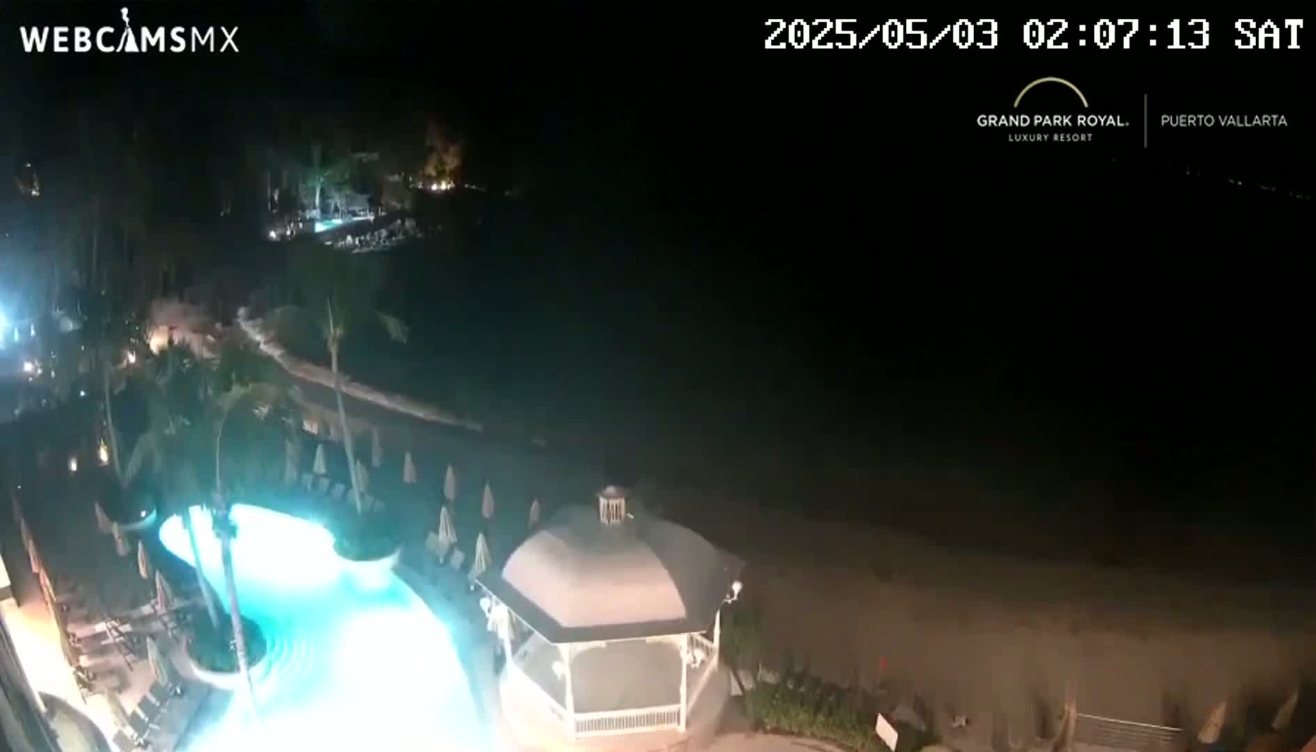 A nighttime view shows a brightly lit swimming pool, a gazebo, and a dark beach under a clear sky.