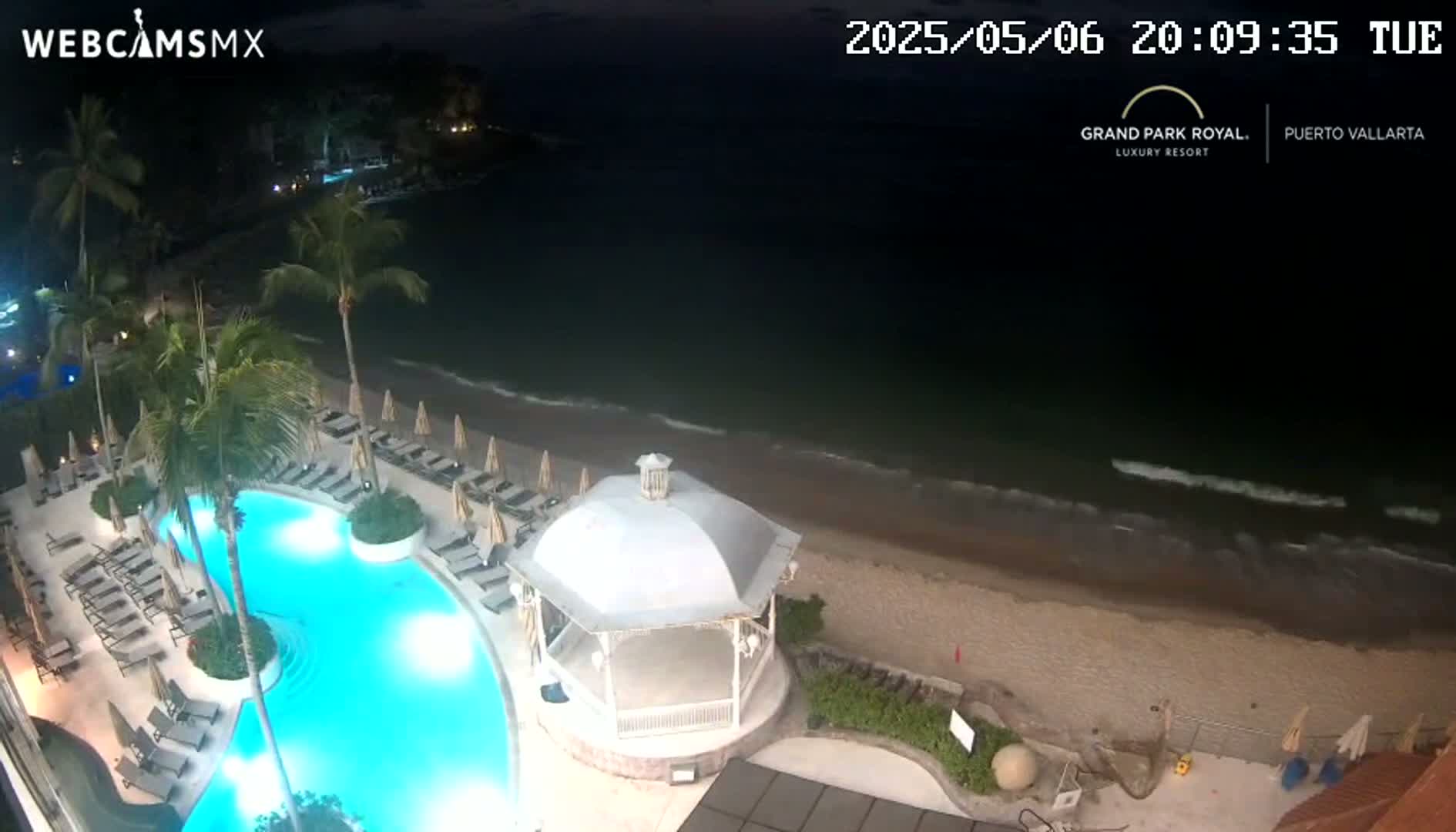 An illuminated resort pool and beach are visible at night, with gentle waves lapping the shore under a dark sky.