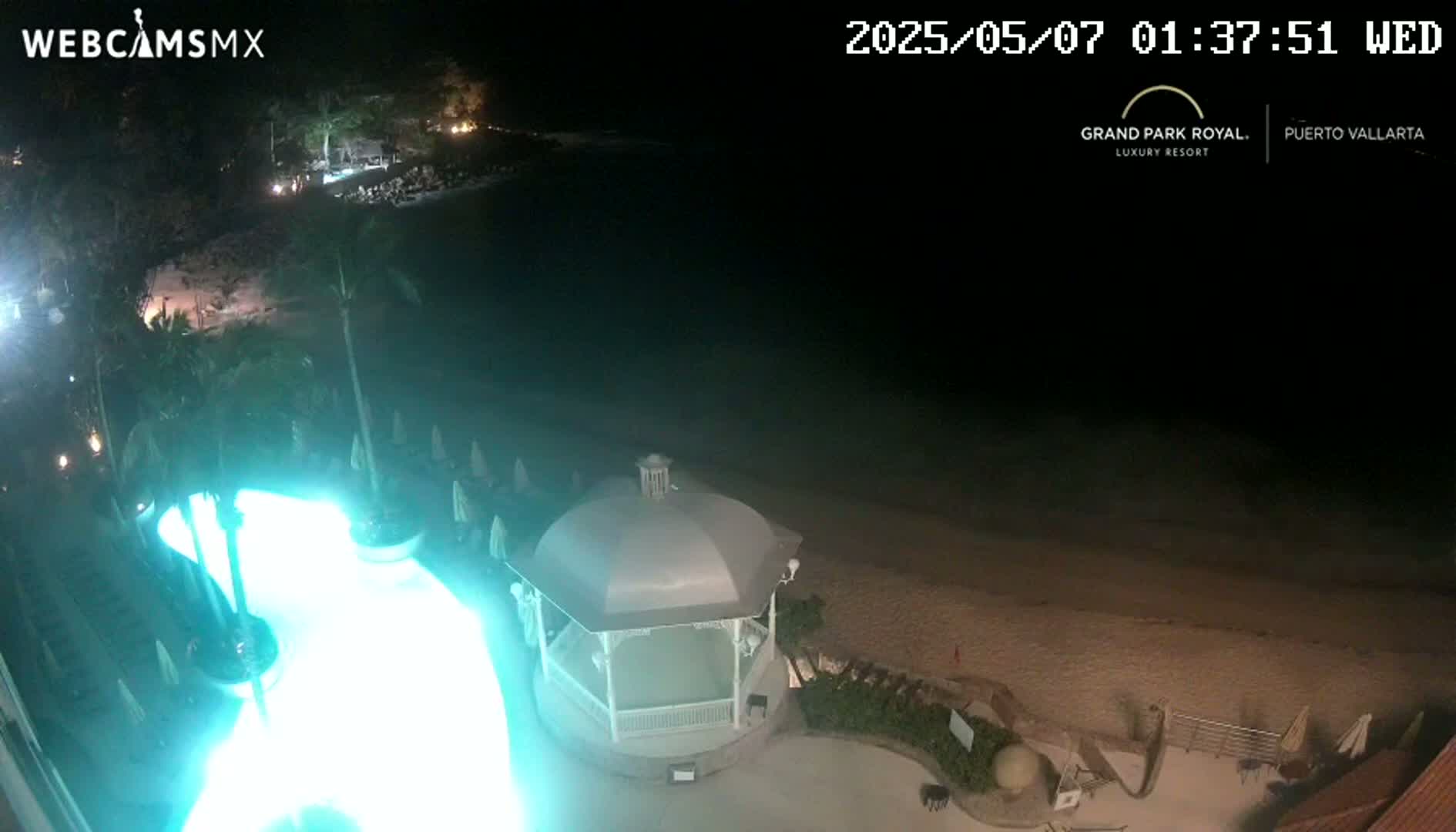 A nighttime aerial view shows a resort pool lit in bright teal, a white gazebo, and a dark sandy beach bordering a calm ocean under a clear, dark sky.