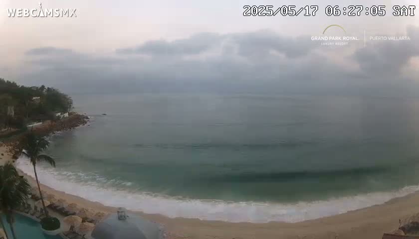 A mostly cloudy day shows a calm ocean with small waves lapping a sandy beach, next to a rocky shore lined with palm trees.
