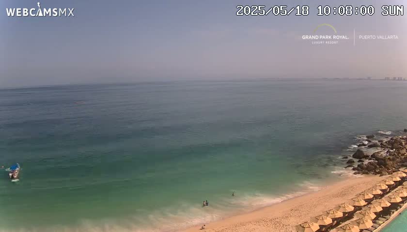 A sunny day shows a tranquil ocean scene with a sandy beach, a few people in the water, a small boat, and beach umbrellas lining the shore.