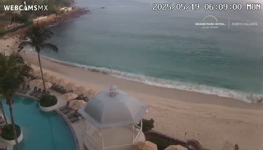An aerial view shows a sandy beach with gentle waves lapping the shore, a swimming pool, and a gazebo next to a resort, under a mostly cloudy sky.
