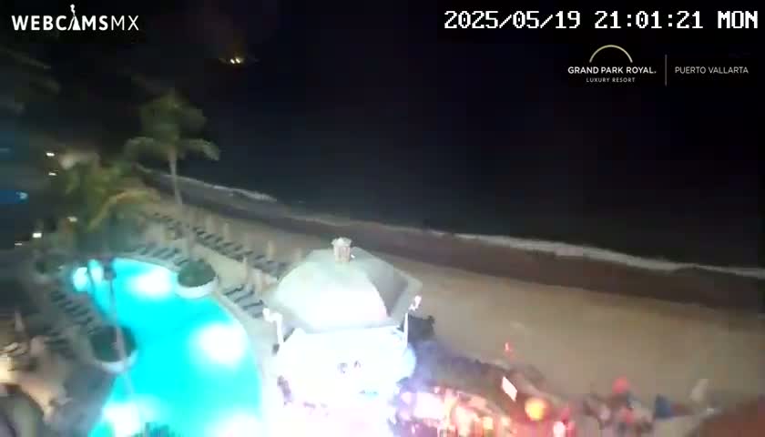 A nighttime aerial view shows a resort pool lit from below, palm trees, a sandy beach with gentle waves, and a gathering of people near the beach.