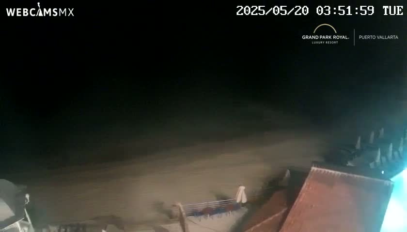 A dark, nighttime beach scene shows a sandy shore and parts of a building under a hazy sky.
