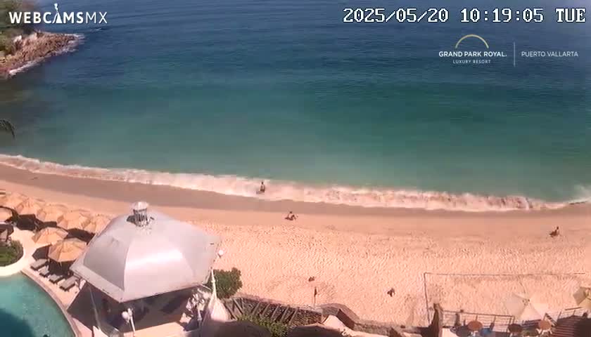 An aerial view shows a sandy beach with small waves lapping the shore on a sunny day, with a few people visible on the beach and in the ocean.