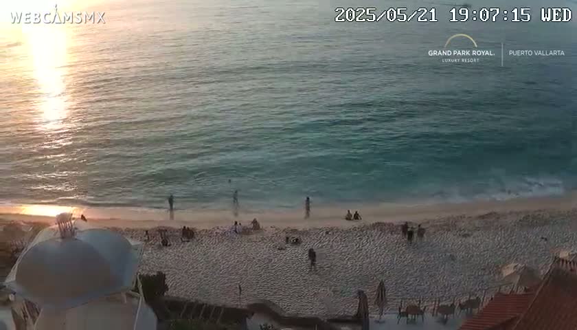 A sandy beach with a few people on it slopes down to a calm, teal ocean reflecting the setting sun.