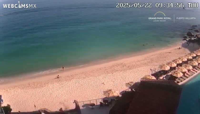 A sandy beach with calm, clear turquoise water and numerous beach umbrellas is visible from an elevated viewpoint on a sunny day.