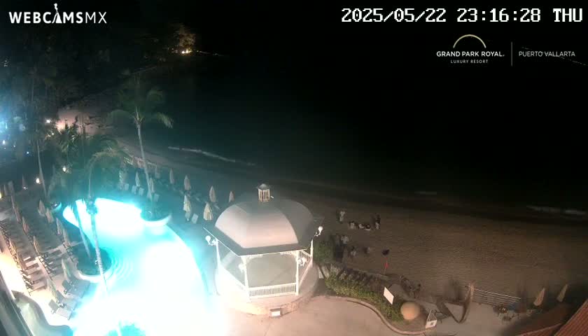 A nighttime aerial view shows a resort pool illuminated in bright teal, a beach, and a gazebo, all under a dark sky.