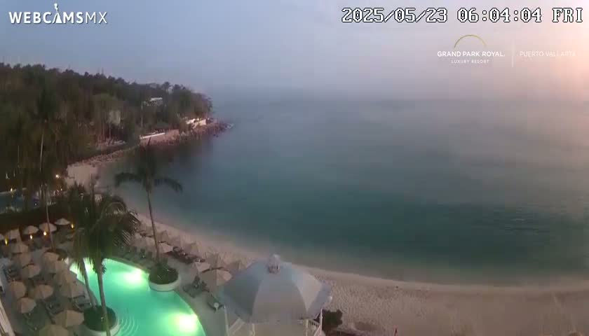 A hazy dawn reveals a resort pool and beach next to a calm ocean, bordered by lush vegetation.