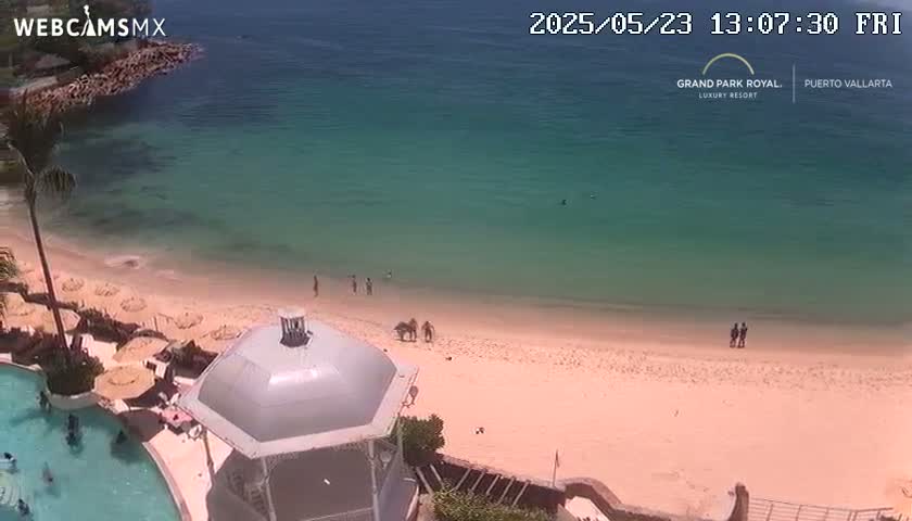 An aerial view shows a sandy beach with calm, clear turquoise water on a sunny day, featuring several people on the beach and in the water near a resort with a gazebo and swimming pool.