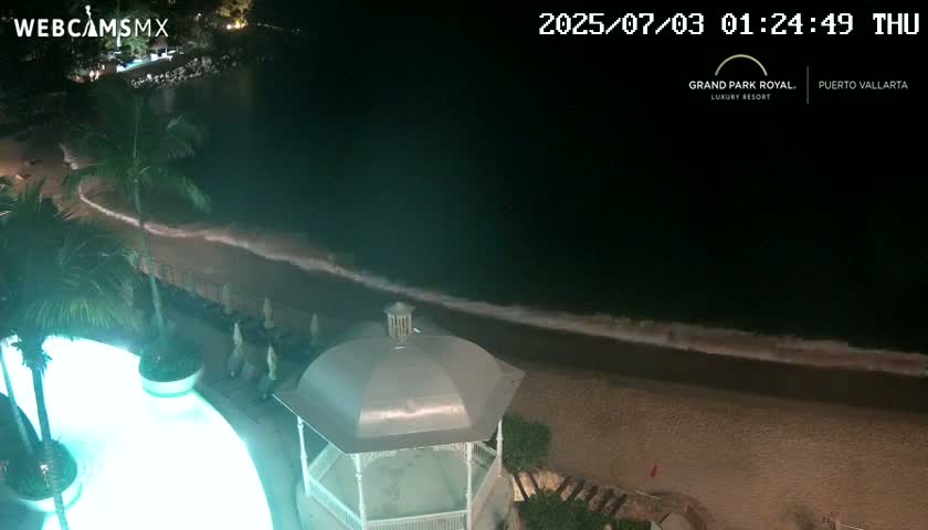 A nighttime view of a resort pool lit in teal, overlooking a sandy beach with gently lapping waves under a dark sky.