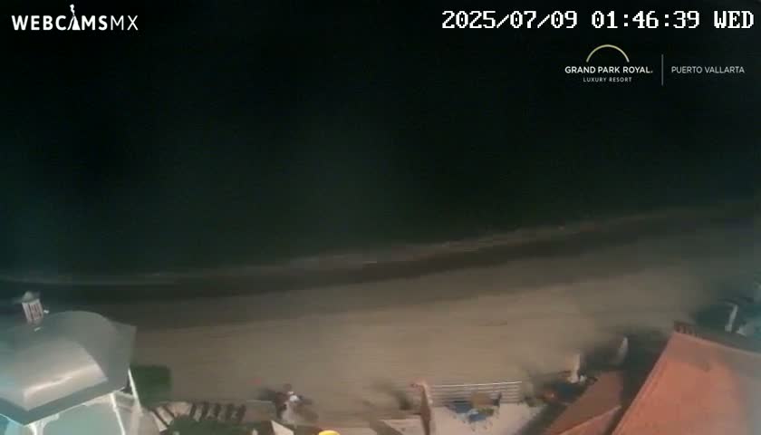 A nighttime, high-angle view shows a dark beach with calm waves and a few people near some buildings.