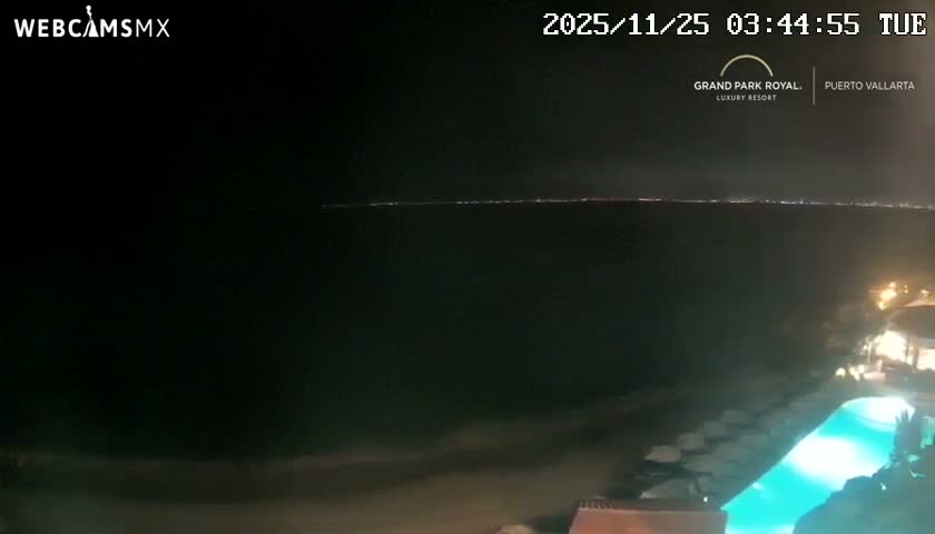 The image shows a clear night view overlooking a body of water with a distant city skyline illuminated across the horizon, while a brightly lit swimming pool and beach chairs are visible in the foreground.