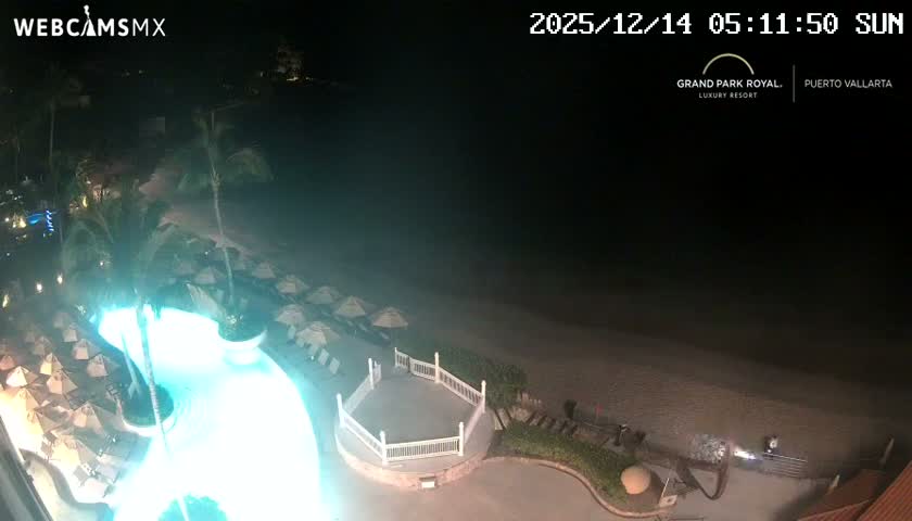 The image shows a clear night view overlooking a body of water with a distant city skyline illuminated across the horizon, while a brightly lit swimming pool and beach chairs are visible in the foreground.