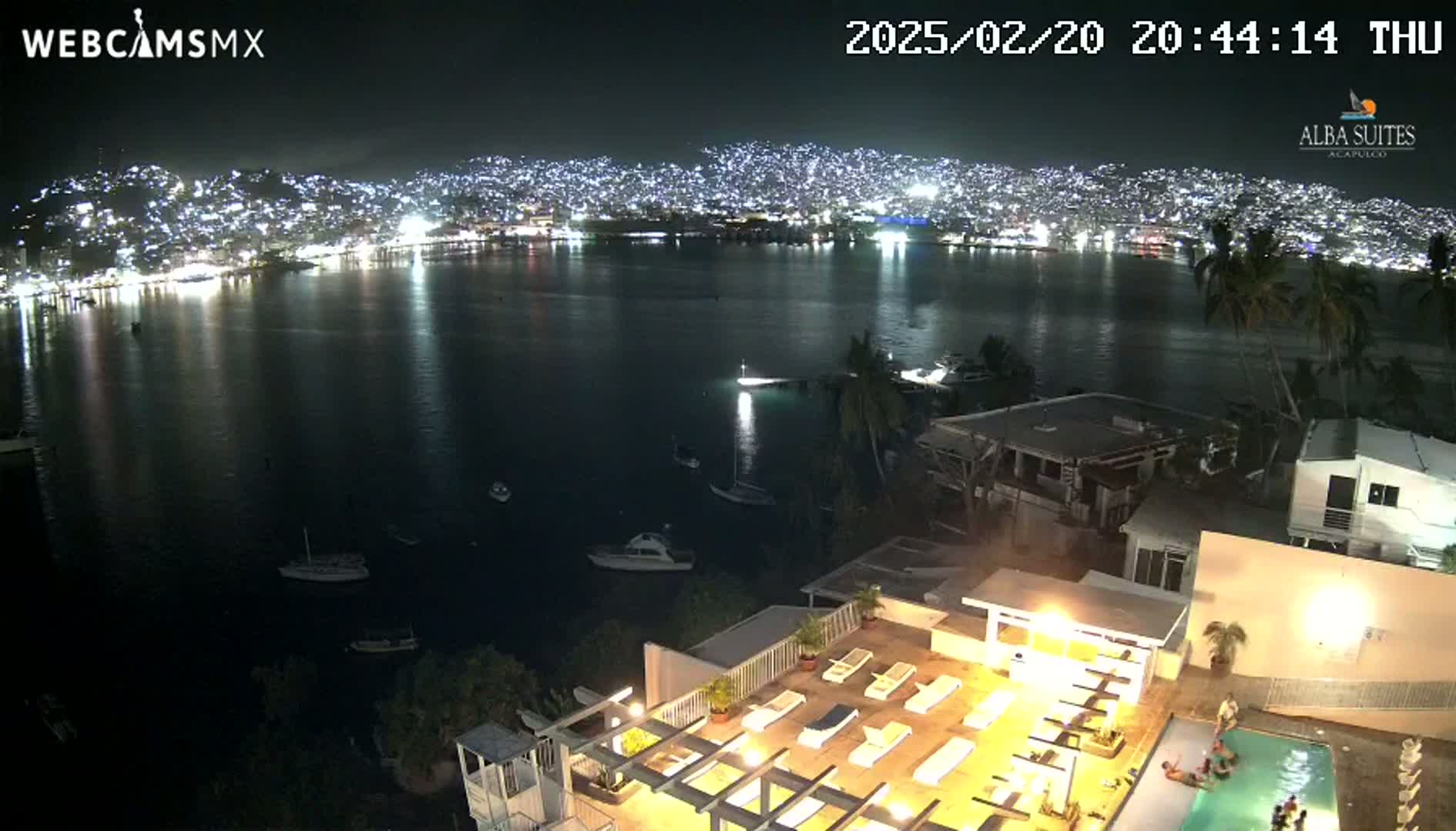 A nighttime view of a city's brightly lit shoreline across a calm bay, seen from a rooftop with a pool and lounge chairs, under a clear sky.