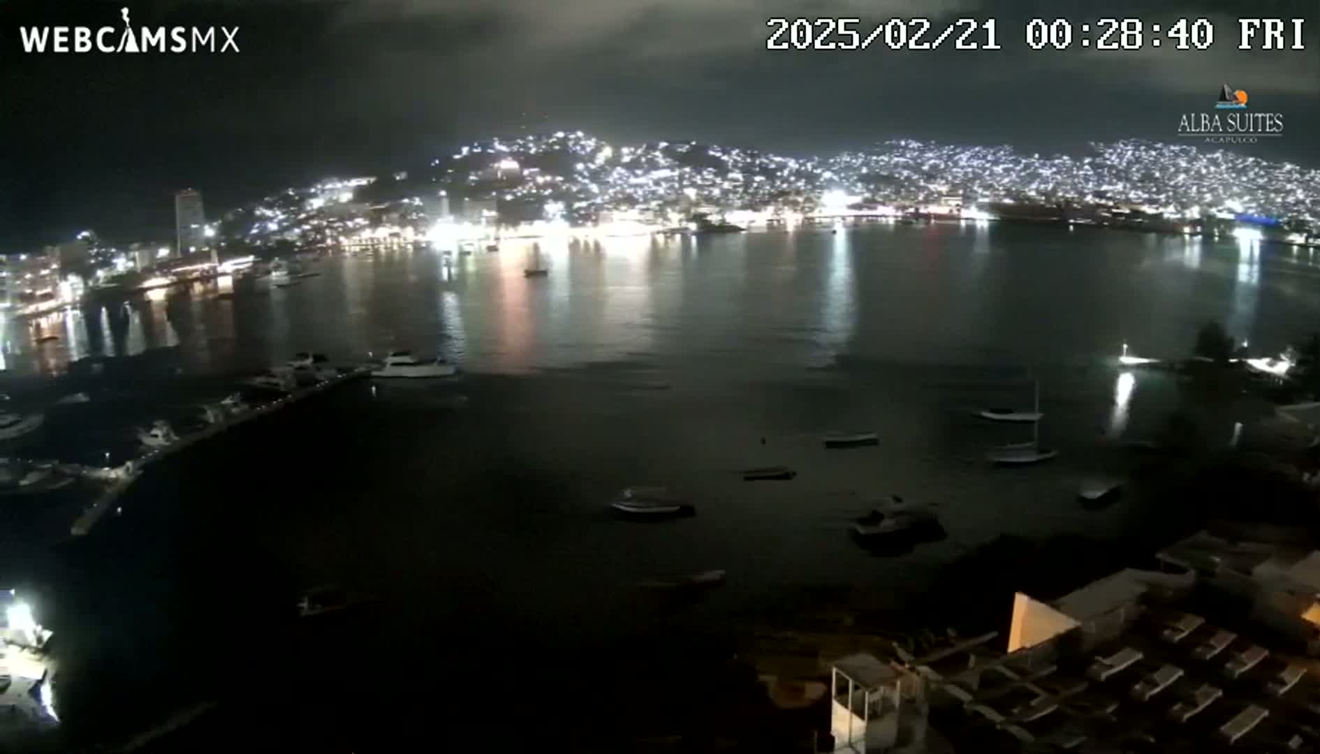 A nighttime view of a city's illuminated coastline, harbor, and docked boats under a dark, cloudy sky.