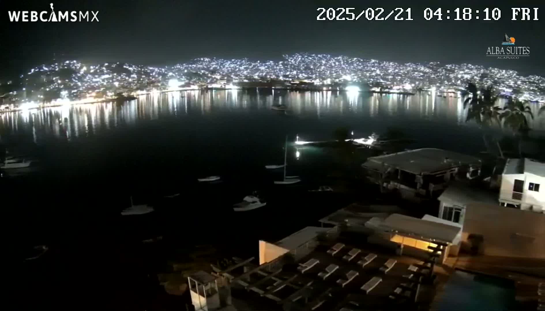 A nighttime view of a city brightly lit across a calm body of water, with several boats and a resort visible in the foreground.