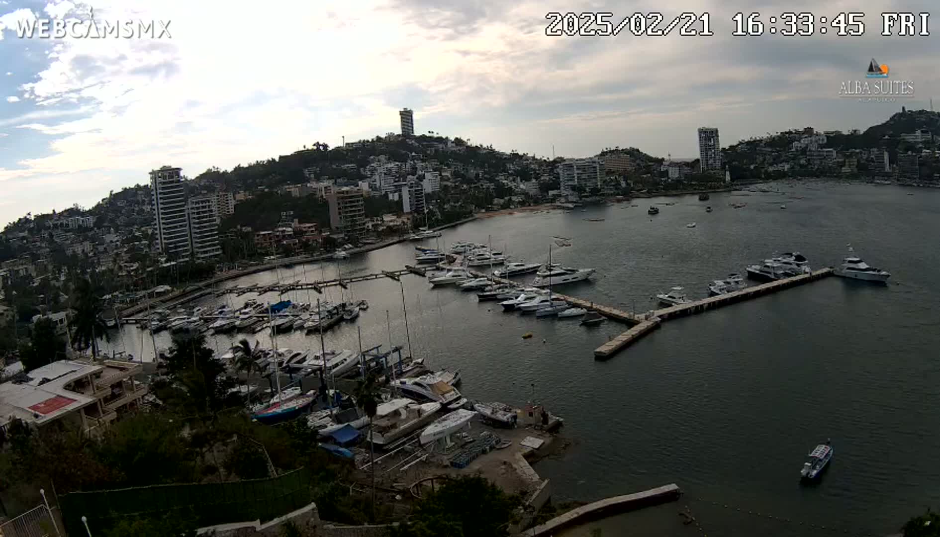 A marina filled with numerous boats and yachts is situated in a bay next to a hillside town under a partly cloudy sky.