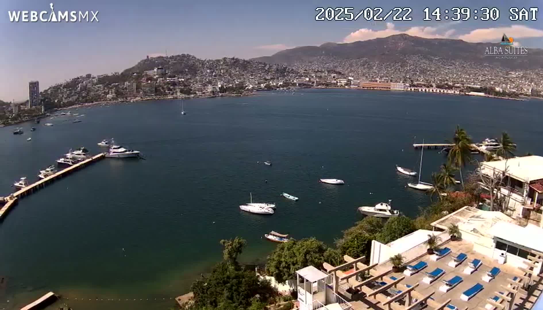 A coastal city on a clear, sunny day is viewed from above, showing a calm bay with several boats and a partially visible resort with lounge chairs.
