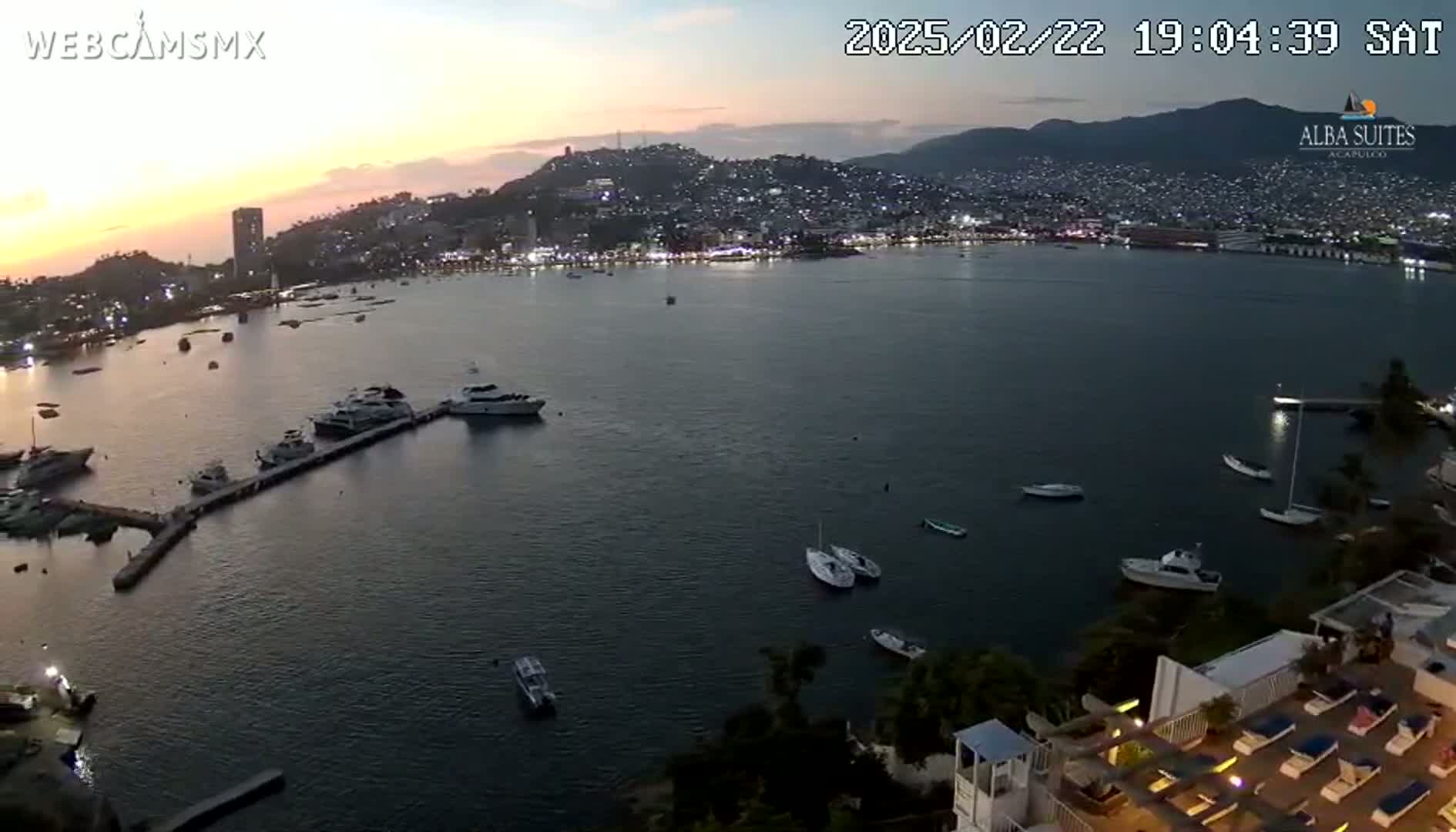 A twilight view of a calm bay with numerous boats docked and anchored, a city skyline in the background, and a rooftop deck with lounge chairs in the foreground.