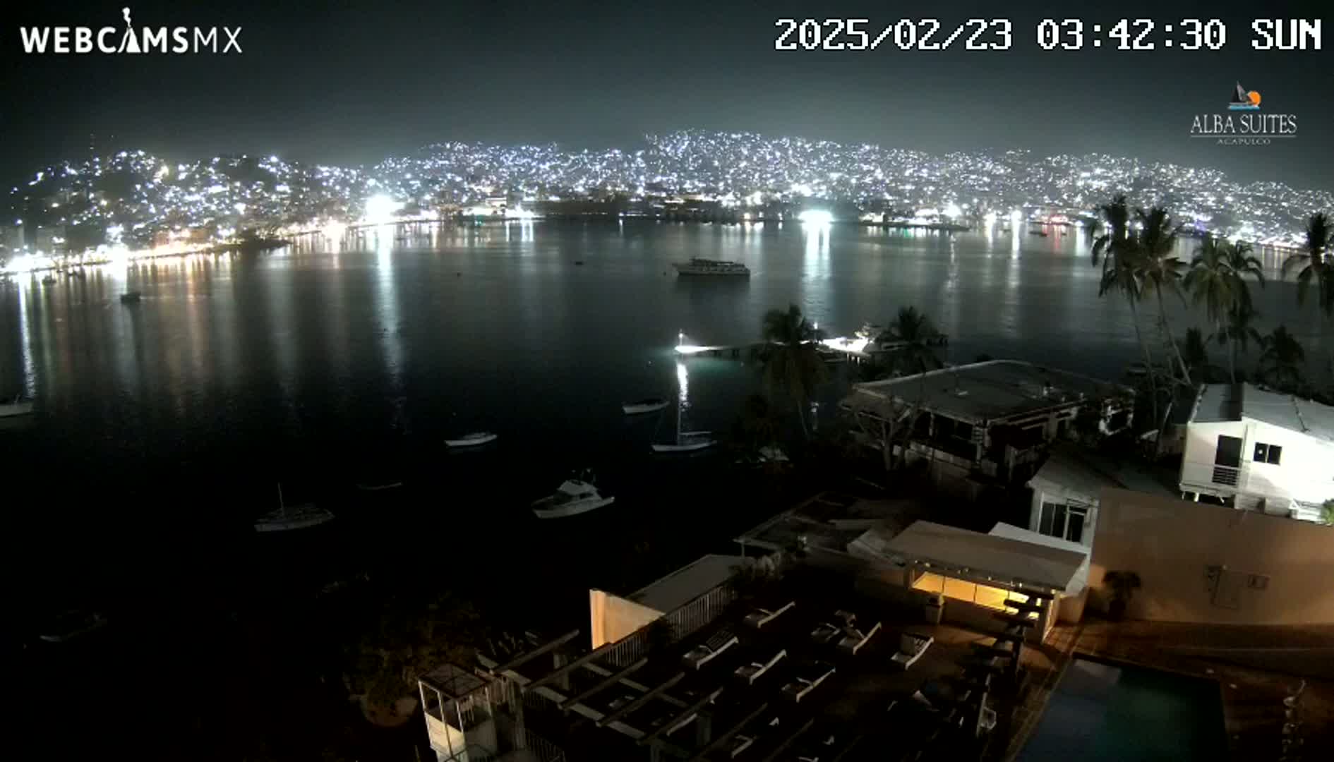 A nighttime view of a city across a calm bay, dotted with boats, seen from an elevated vantage point overlooking a building with a pool.