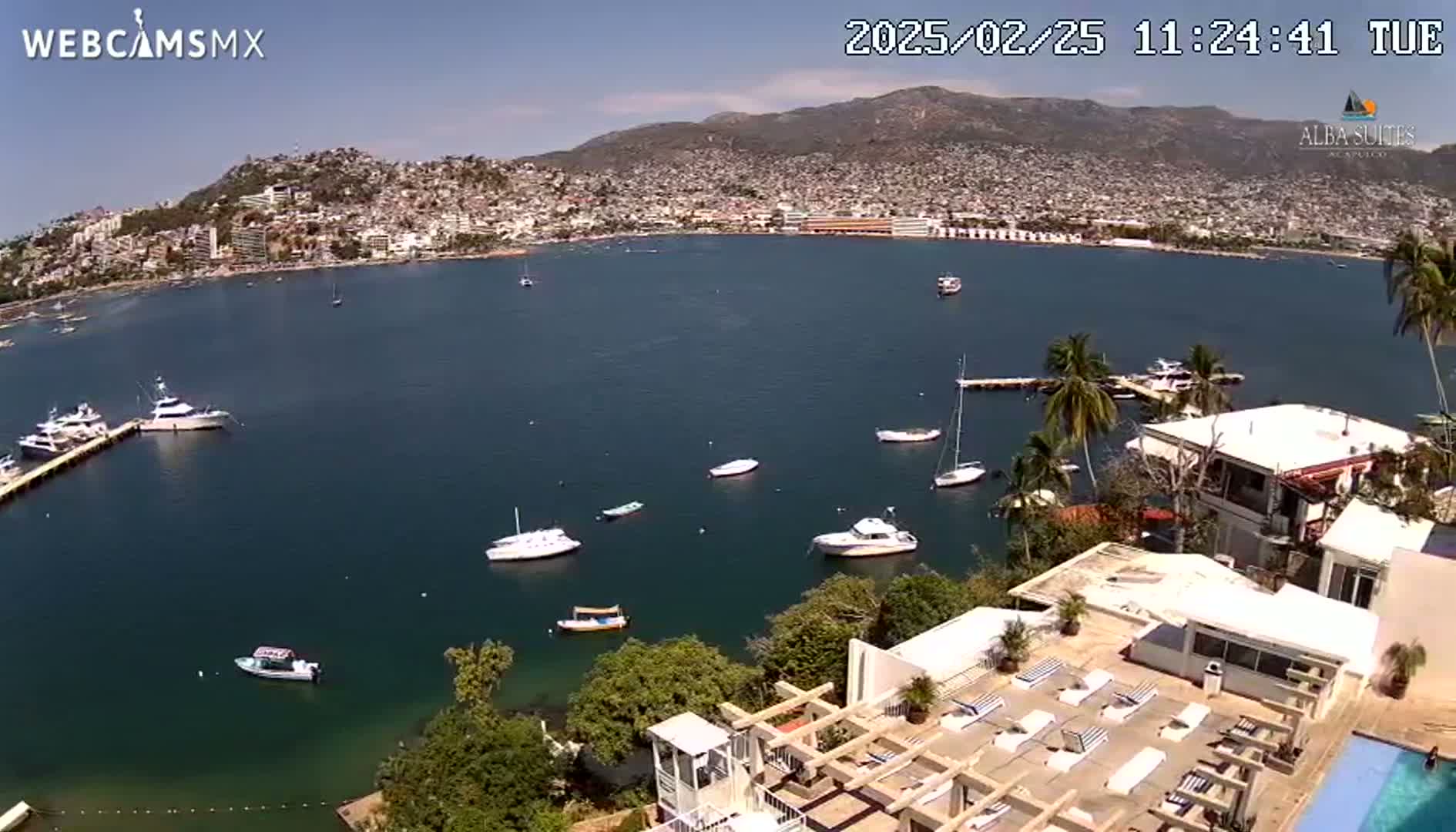 An aerial view shows a calm bay with numerous boats, a hillside city, and mountains under a clear, sunny sky.