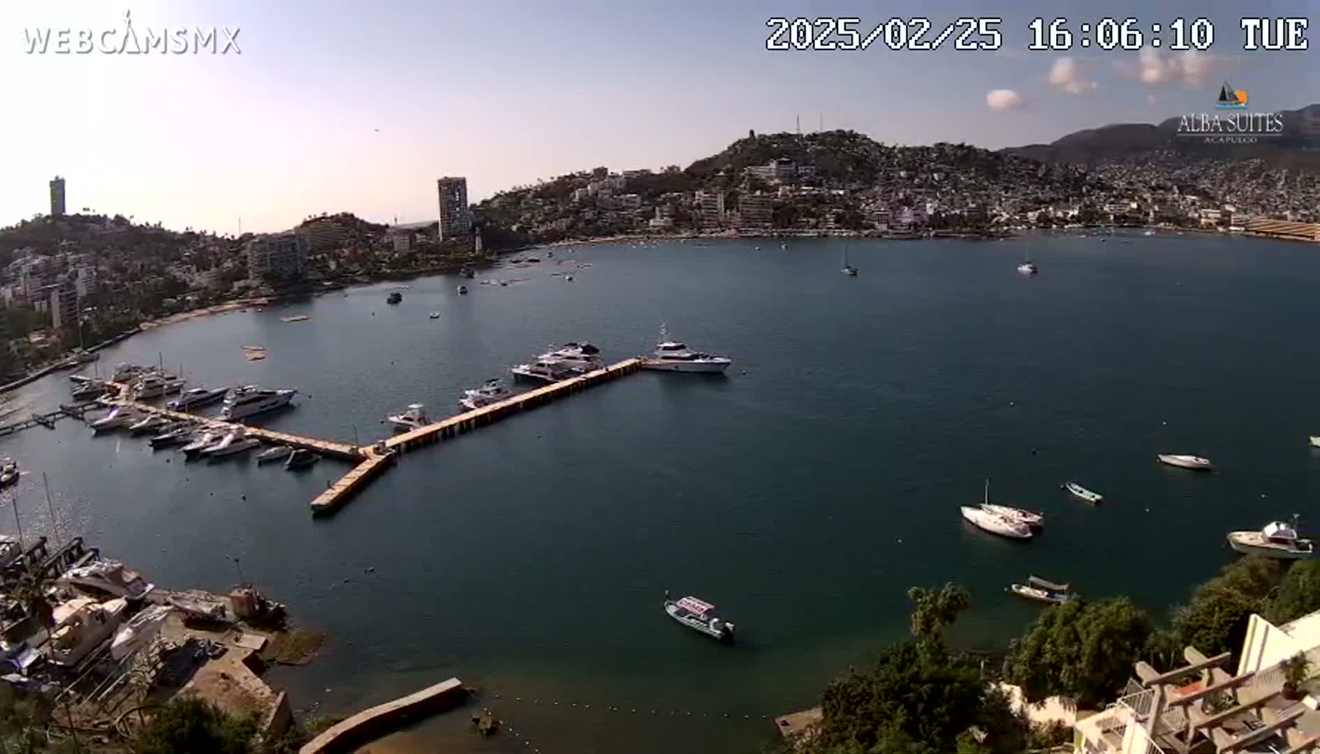 A calm bay with numerous boats docked at a pier, surrounded by a hillside town under a mostly clear sky.