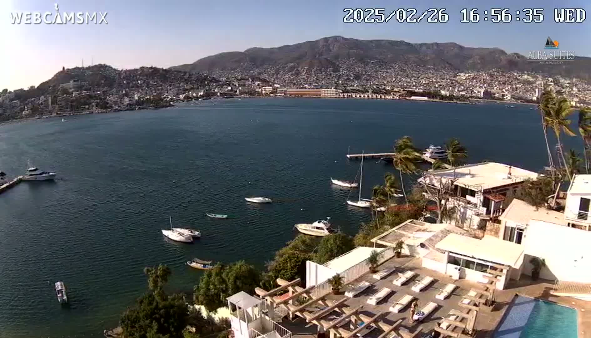 An aerial view shows a calm bay with several boats, a city nestled against mountains in the background, and a resort with a pool and sun loungers in the foreground under a clear, sunny sky.