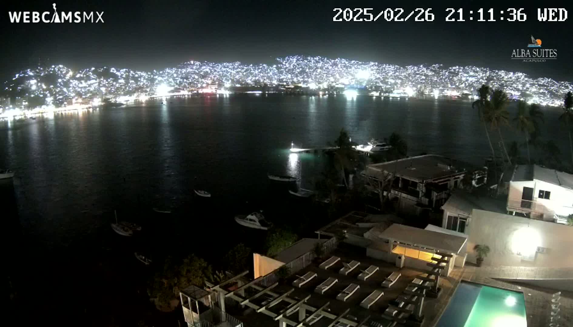A nighttime view from a high vantage point overlooking a calm bay with boats, a brightly lit city on the opposite shore, and a swimming pool at a building in the foreground.