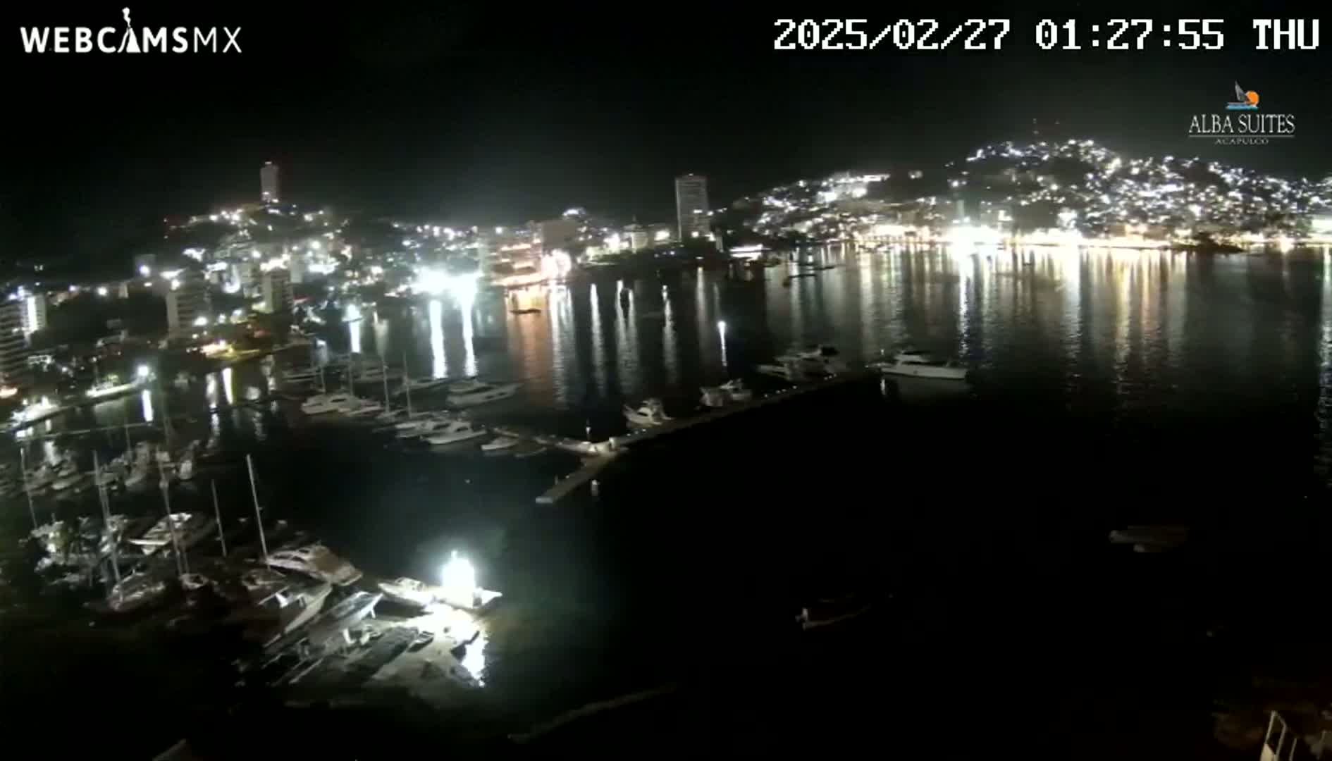 A nighttime view of a harbor filled with boats, reflecting the lights of a city on a calm body of water.
