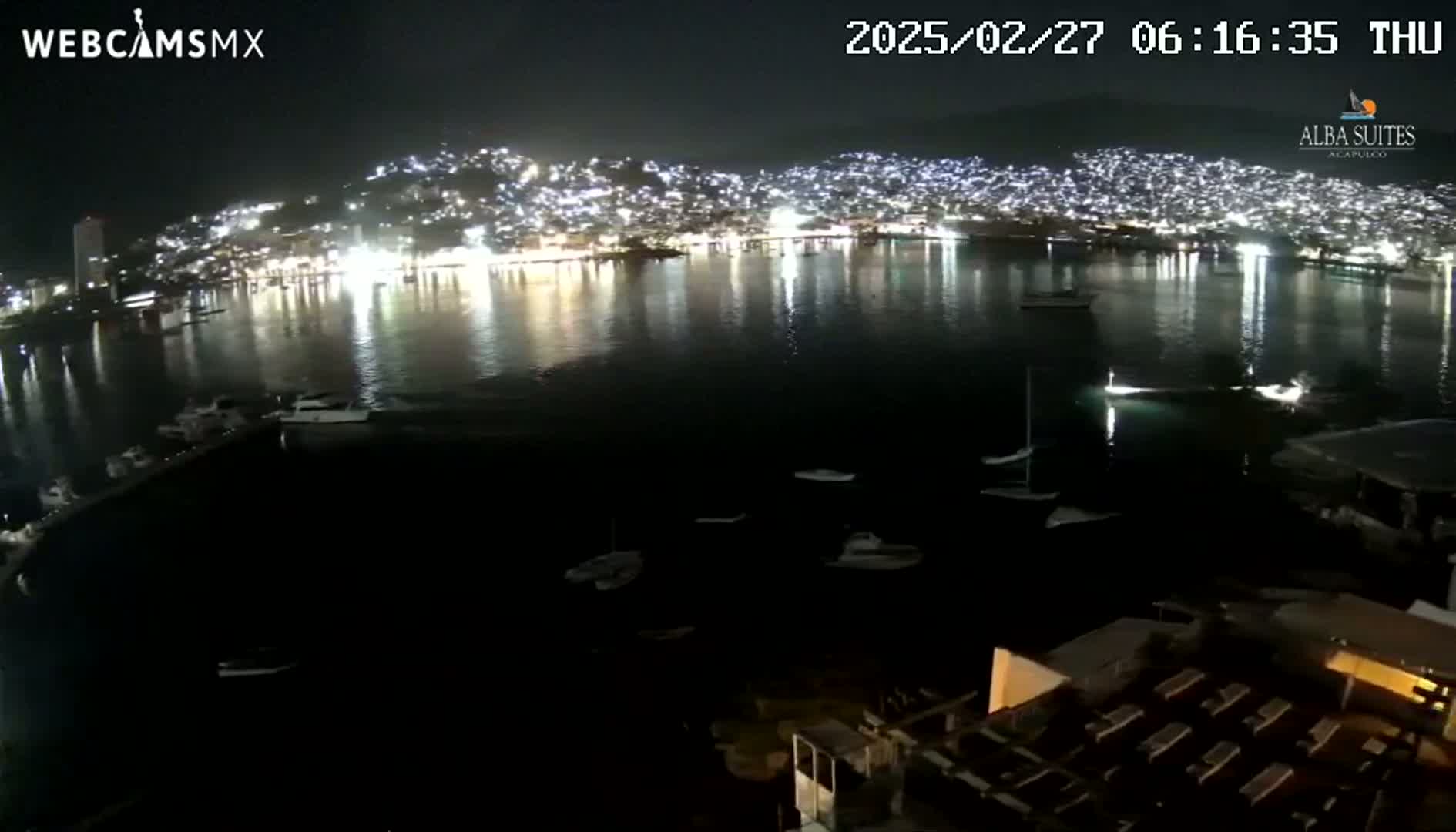 A brightly lit city on a hillside is reflected in a dark body of water at night, with several boats visible in the harbor.