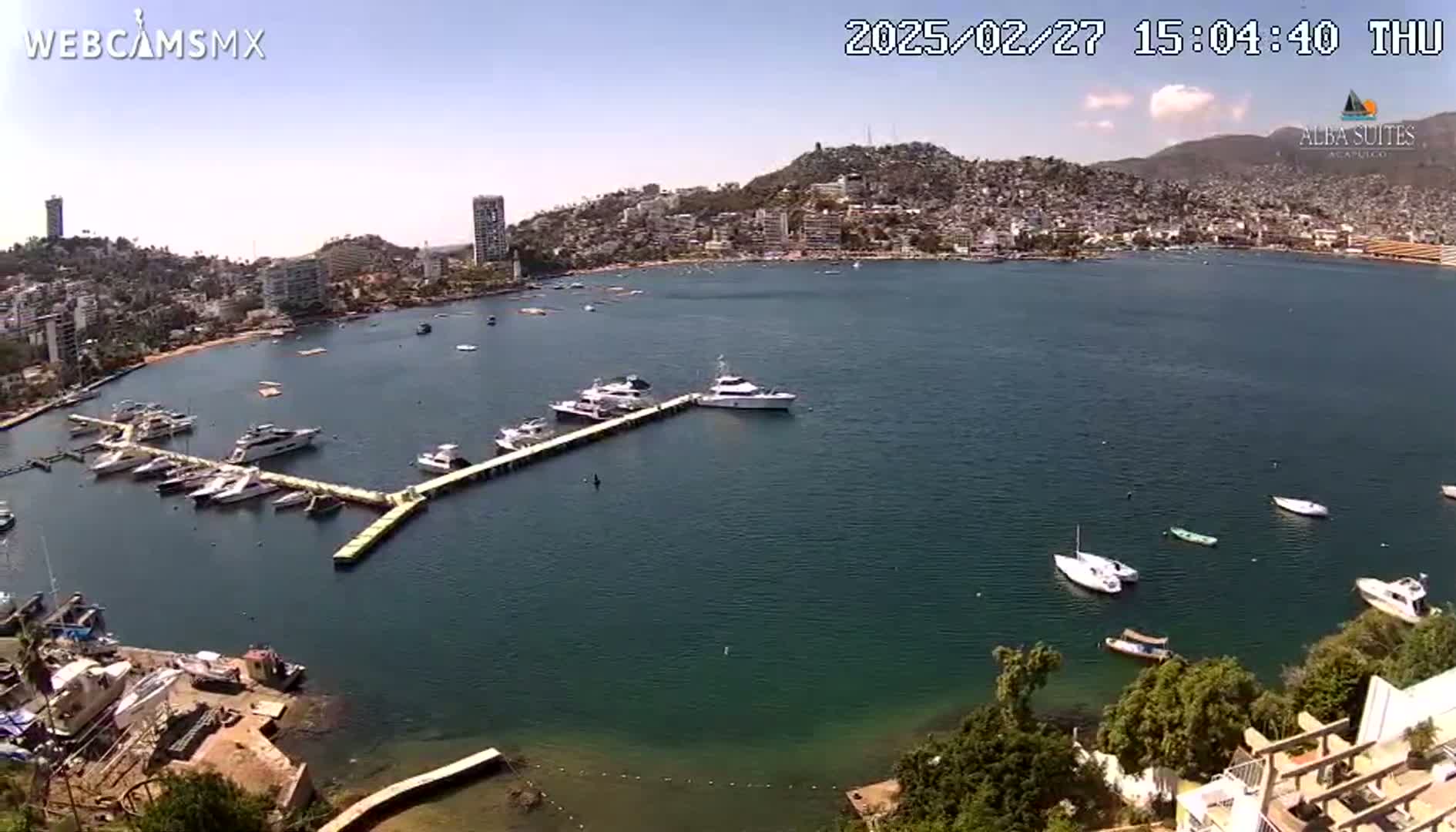 A calm bay with numerous boats docked at a pier, surrounded by a city nestled against a hillside under a clear, sunny sky.
