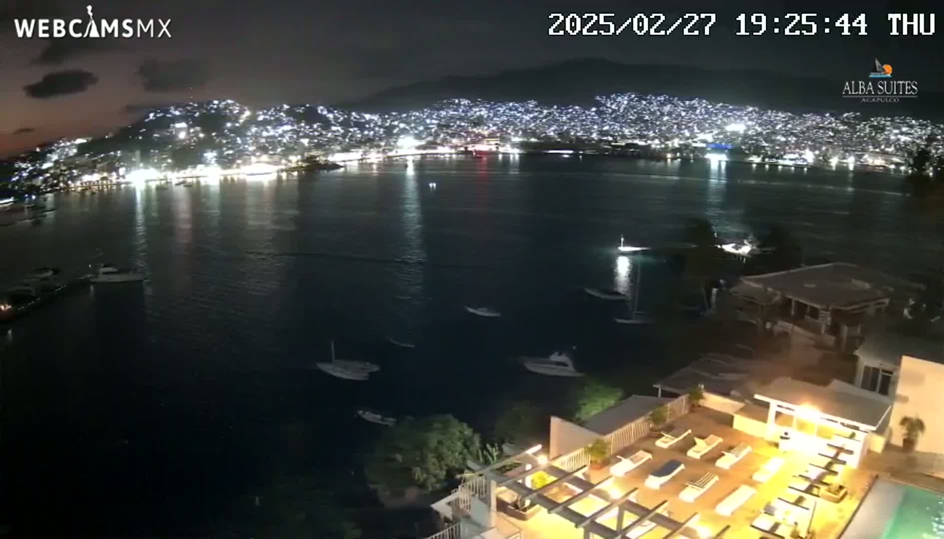 A nighttime view of a city illuminated across a dark bay, seen from a high vantage point overlooking a rooftop terrace with lounge chairs and a pool.