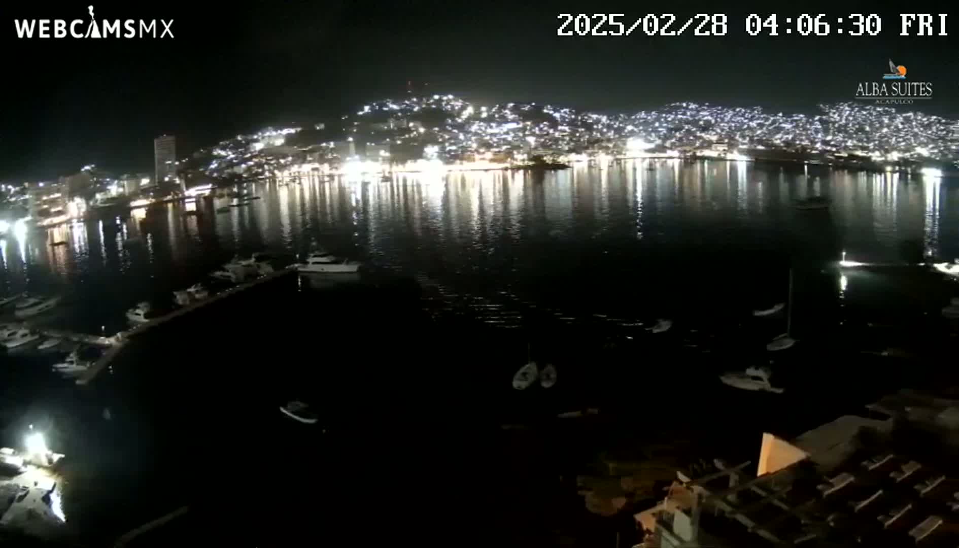 A nighttime view of a city harbor with numerous boats docked and many city lights reflecting on calm water.