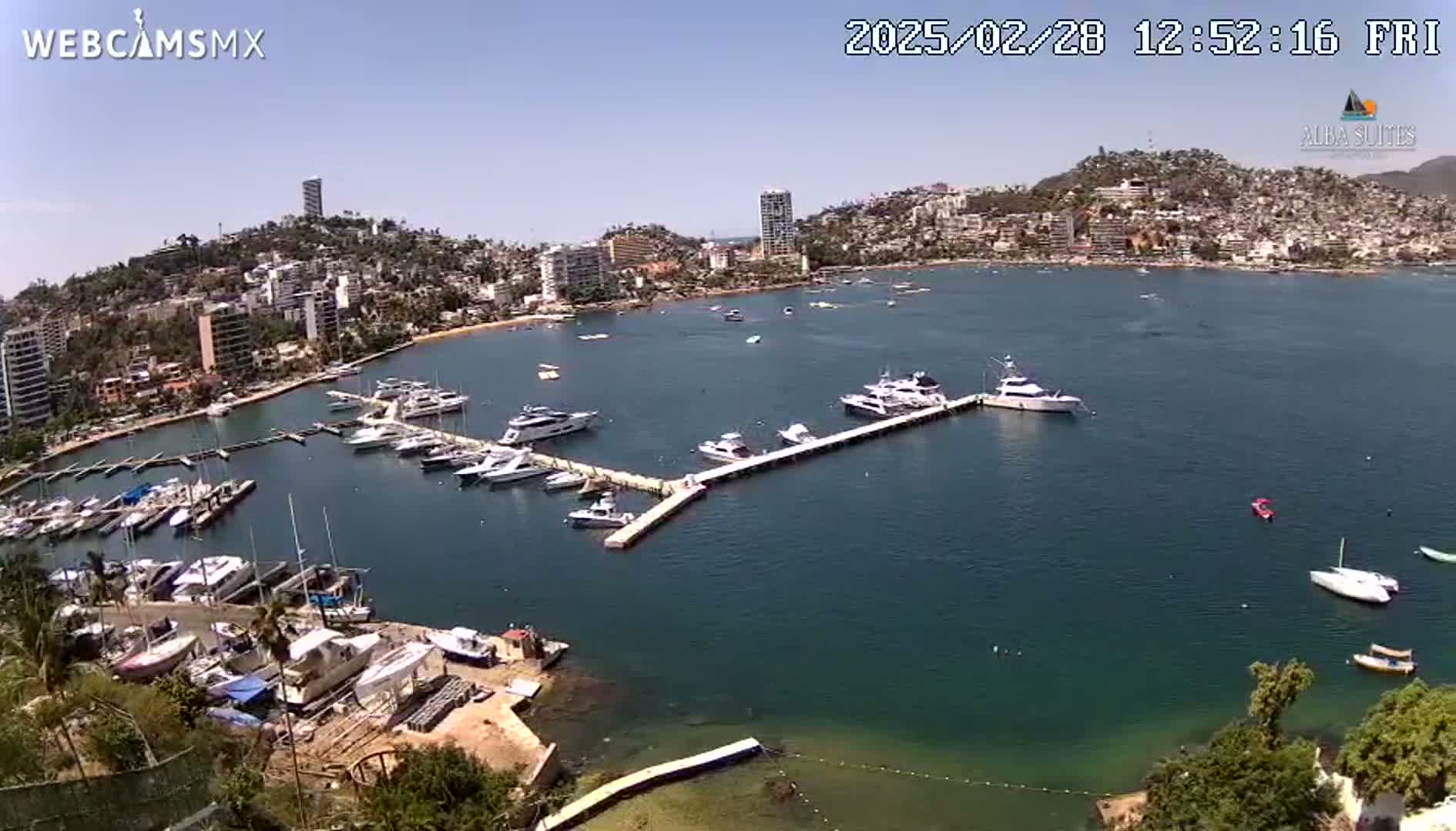 A sunny aerial view shows a marina filled with boats in a calm bay next to a city built on a hillside.