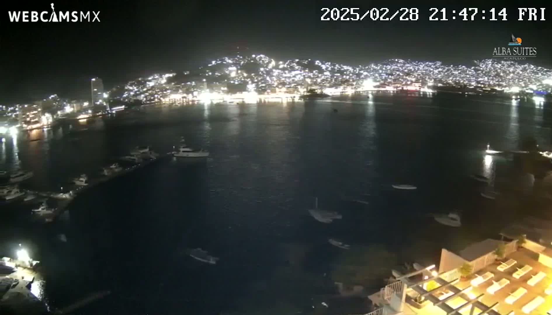 A nighttime view of a city illuminated along a calm bay, dotted with boats.