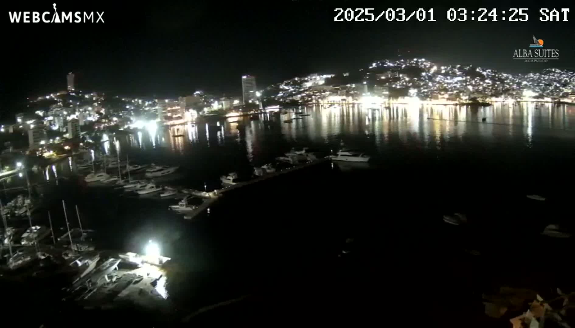 A nighttime view of a harbor filled with boats, reflecting the lights of a brightly lit city on the hills behind it.