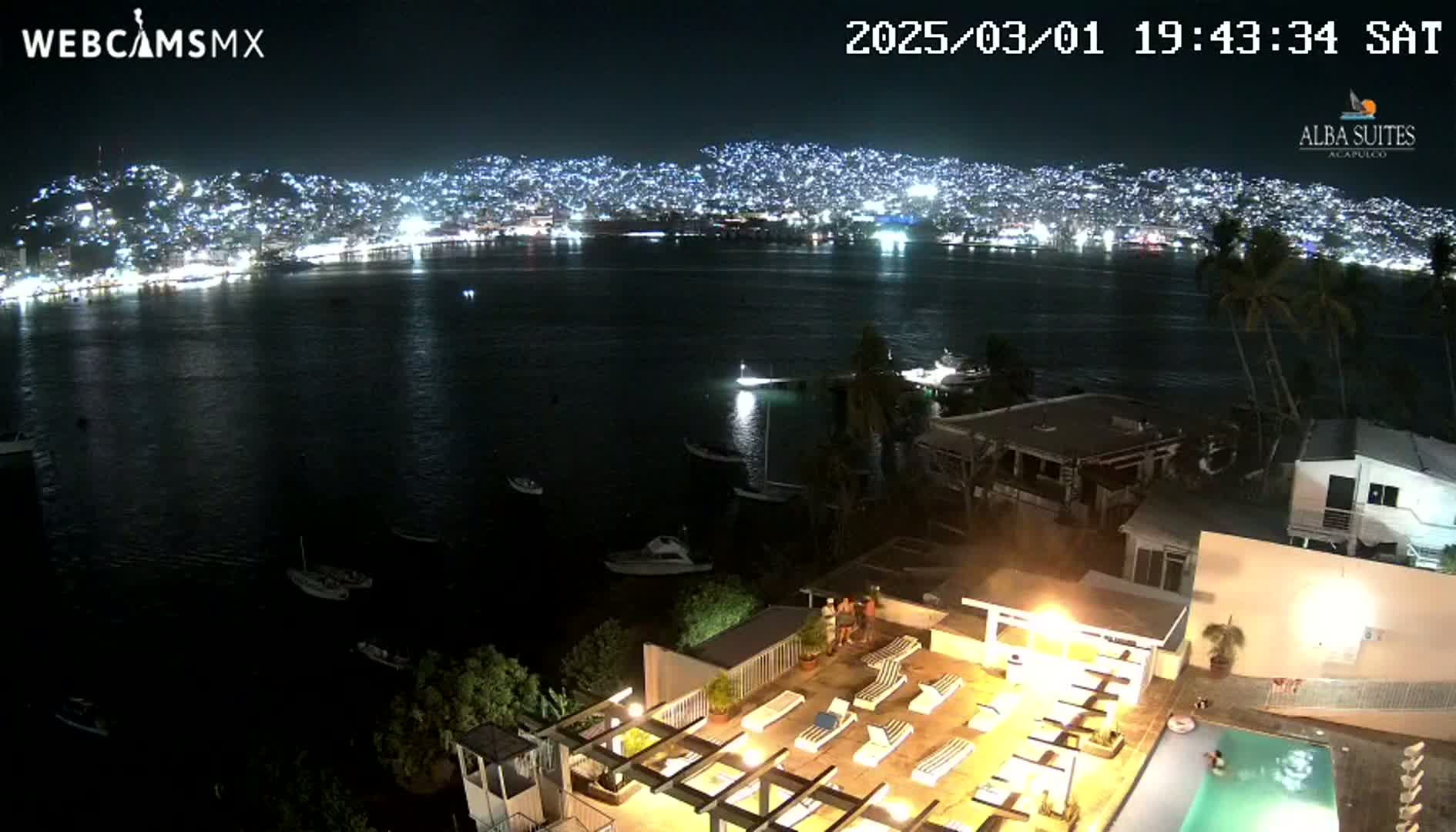 A nighttime view of a city illuminated across a dark bay, seen from a rooftop with a pool and lounge chairs, under a clear sky.