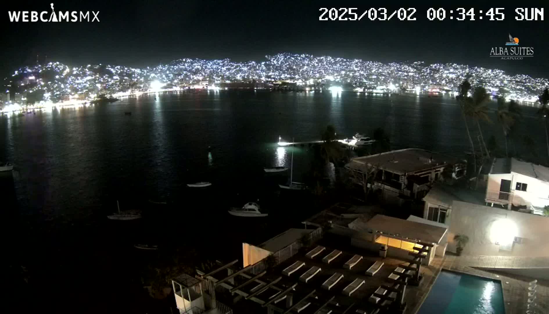 A nighttime view of a city's illuminated coastline across a dark body of water, with several boats visible, and a building with a pool in the foreground.
